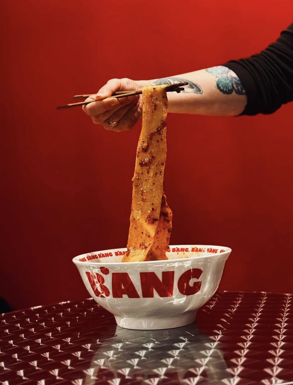 A wide biang biang noodle lifted with chopsticks from a BANG BANG bowl