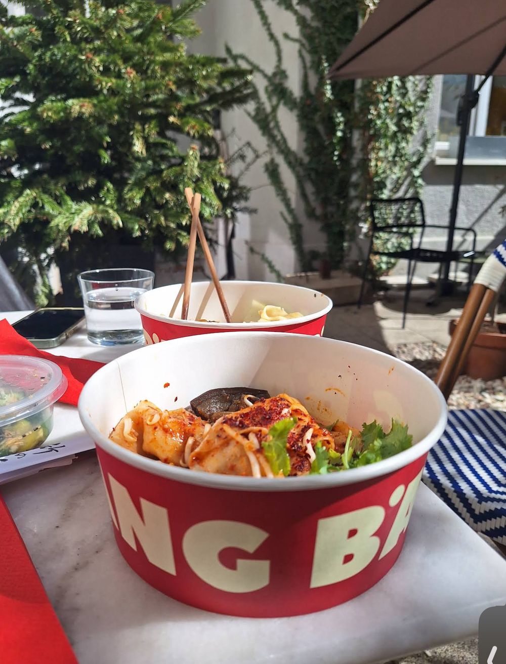 A red BANG BANG takeaway cup of noodles on a sunny patio table